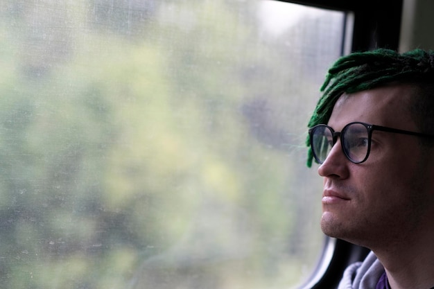 Portrait of young man with green dreadlocks in glasses riding on train looking at nature Close up