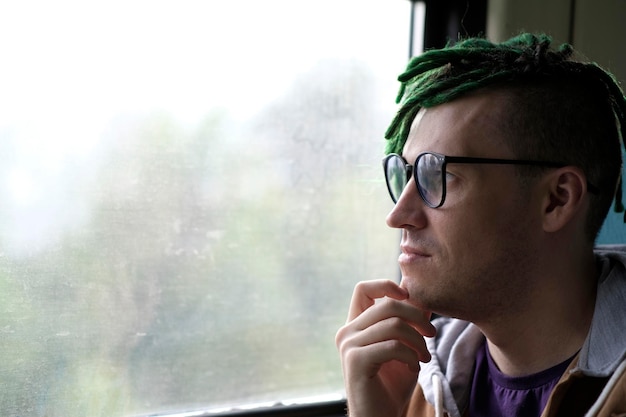 Portrait of young man with green dreadlocks in glasses riding on train looking at nature Close up