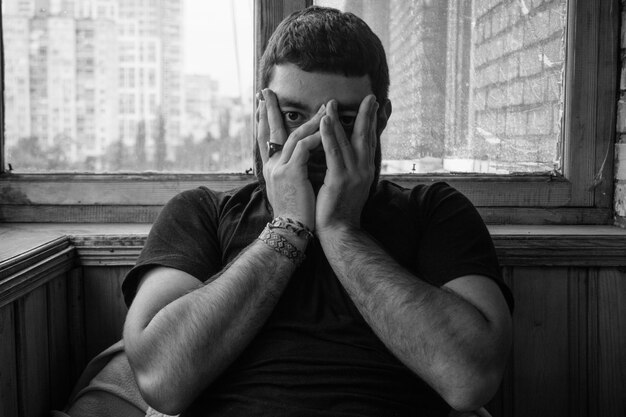 Photo portrait of young man with hands covering mouth sitting against window at home