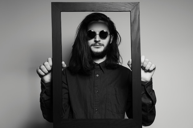 Portrait of young man with long hair holding a dark frame close to his face
