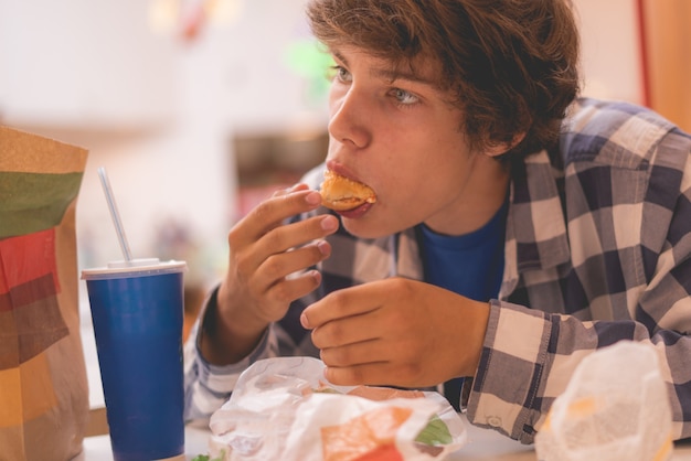 Portrait of young man with long hair sitting at the table and eating and biting hamburger