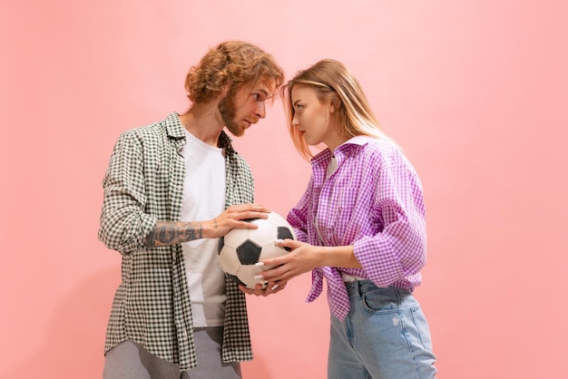 Portrait of young man and woman football fans arguing posing isolated over pink studio background