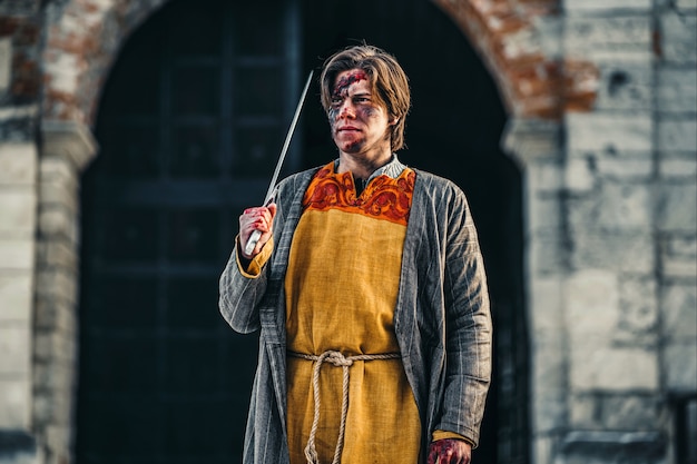 Portrait of a young medieval warrior in armor with sword on his shoulder and wounds on the face. In the process of battle, near the castle