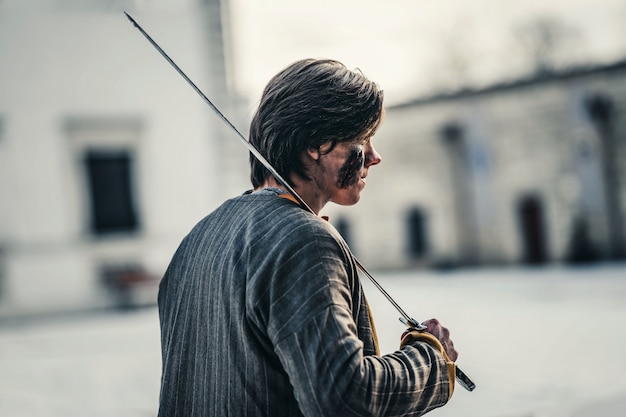 彼の肩に剣と顔に傷を持つ鎧を着た若い中世の戦士の肖像画。戦いの最中、城の近く