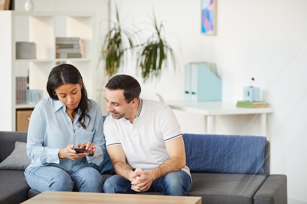 Portrait of young mixed-race woman showing smartphone to male colleague while sitting on couch in office interior