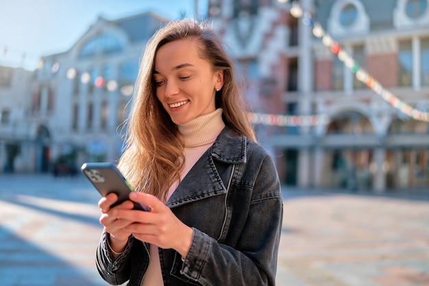 Ritratto di giovane donna allegra felice caucasica attraente casuale moderna che tiene e che utilizza telefono all'aperto