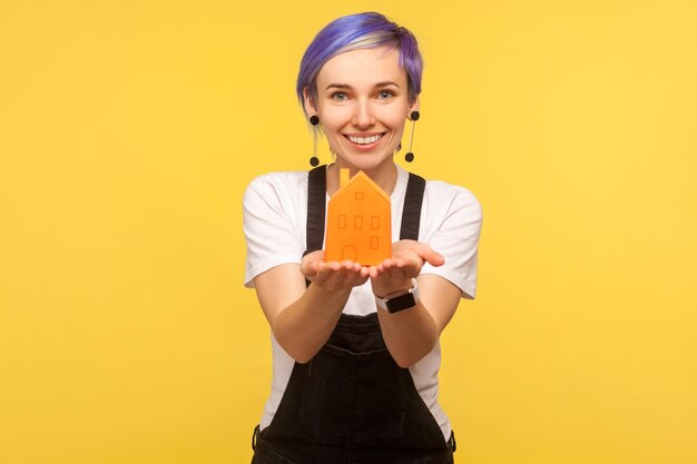 Portrait of young modern fashionable hipster girl with violet short hair in denim overalls holding orange paper house on palms and smiling at camera indoor studio shot isolated on yellow background