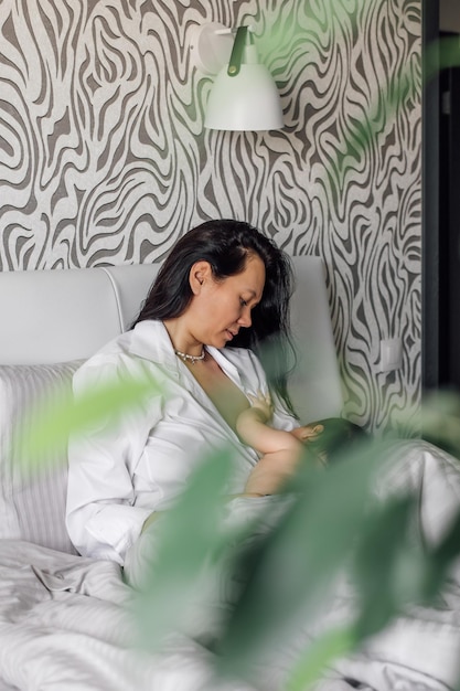 Portrait of young mother breastfeeding her daughter in bedroom blurred green plant in foreground Baby suckle milk and get nutrients for healthy growth Concept of maternal affection and childcare