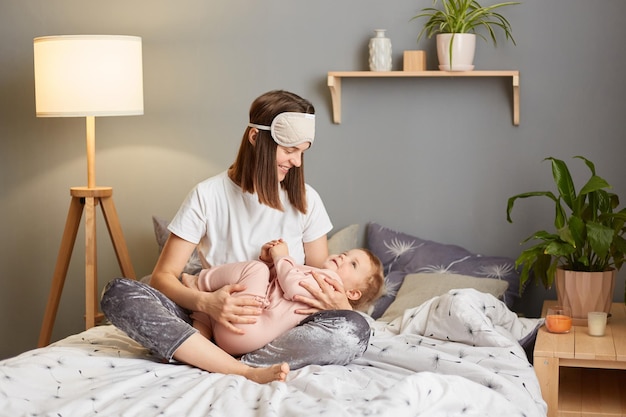 Portrait of young mother in sleep mask spending time with her baby daughter child embracing and hugging kid while sitting on the bed mommy lulling her toddler infant child