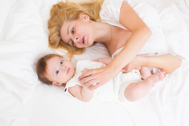 Ritratto di giovane madre con capelli biondi con la sua dolce bambina di 3 mesi in abito bianco che si diverte in camera da letto al mattino, amorevole concetto di famiglia felice, vista dall'alto