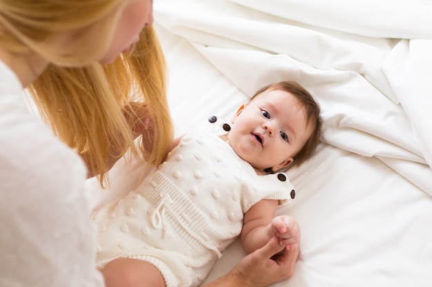 Portrait of young mother with blond hair with her sweet 3 month old baby girl in white wear having fun in the bedroom at morning, loving happy family concept