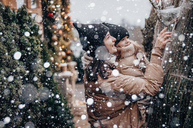 Portrait of a young mother with daughter in her arms they are smiling and rejoicing in the snowfall Choosing and buying a Christmas tree at the Christmas market
