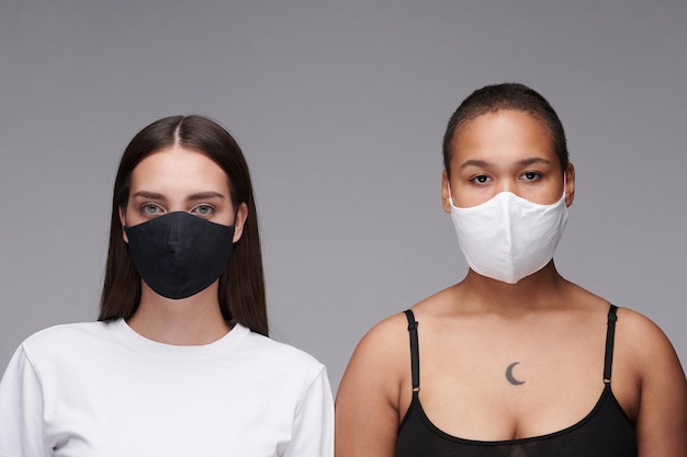 Portrait of young multiethnic women in black and white masks standing against gray background coronavirus concept