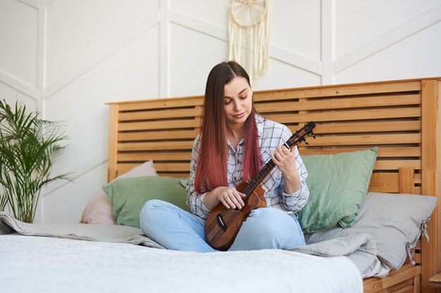 Foto ritratto di un giovane musicista che suona l'ukulele seduto sul letto di casa sua, a gambe incrociate.