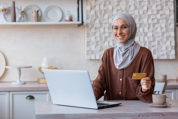 Portrait of young muslim business woman in hijab sitting at home at table with laptop has a credit