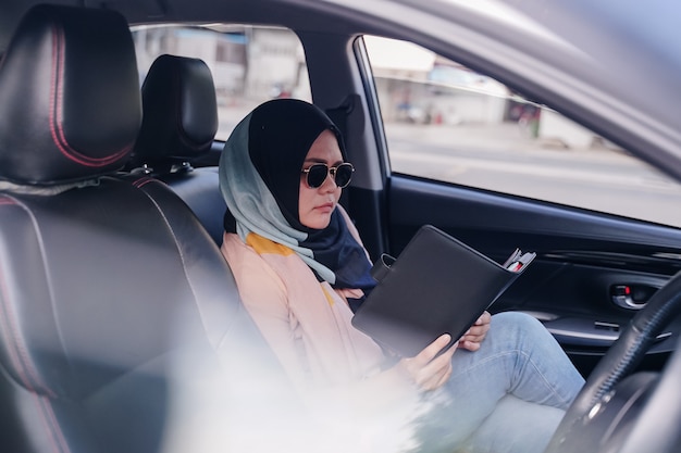 Photo portrait of a young muslim business woman reading in the back seat of the car.