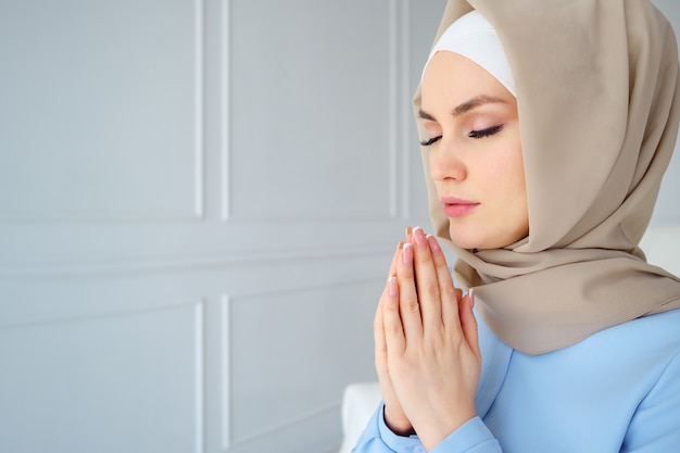 Portrait of young muslim woman in beige hijab and traditional clothes praying with eyes closed, copy space.