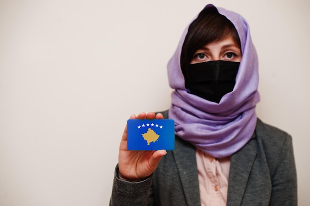 Portrait of young muslim woman wearing formal wear protect face mask and hijab head scarf hold Kosovo flag card against isolated background Coronavirus country concept