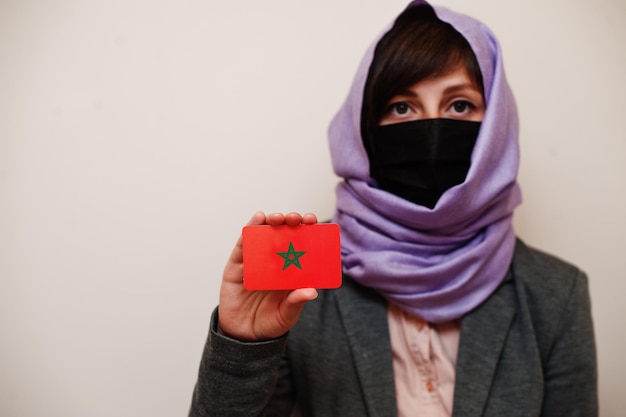 Portrait of young muslim woman wearing formal wear protect face mask and hijab head scarf hold Morocco flag card against isolated background Coronavirus country concept