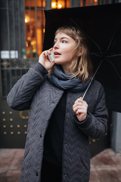 Portrait of young nice lady standing on street with black umbrella and talking on her cellphone