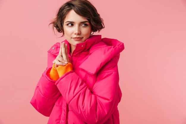 Portrait of young nice woman in warm coat making gun gesture and looking aside isolated on pink