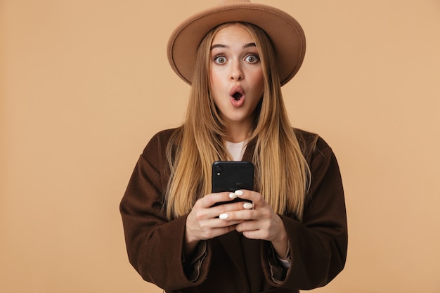 Portrait of young optimistic girl wearing hat wondering and holding cellphone isolated on beige