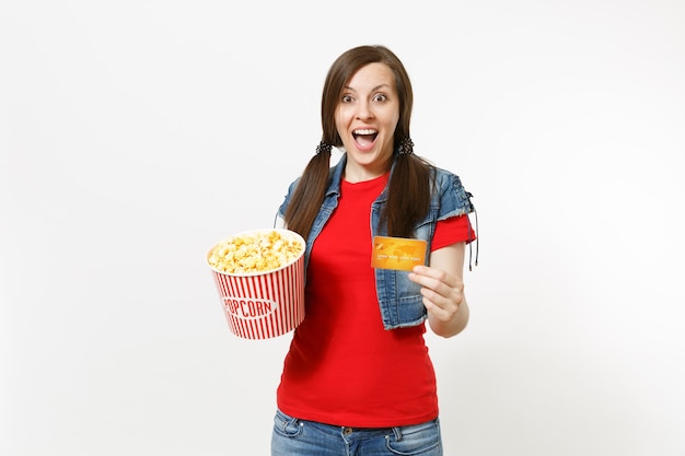 Portrait of young overjoyed attractive brunette woman in casual clothes watching movie film, holding bucket of popcorn and credit card isolated on white background. Emotions in cinema concept.