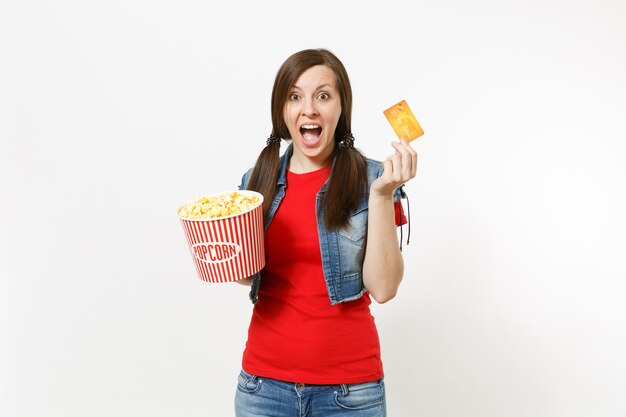 Portrait of young overjoyed attractive brunette woman in casual clothes watching movie film, holding bucket of popcorn and credit card isolated on white background. Emotions in cinema concept.