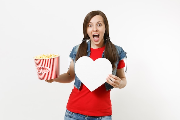 Portrait of young overjoyed shocked woman in casual clothes watching movie film, holding bucket of popcorn and white heart with copy space isolated on white background. Emotions in cinema concept.