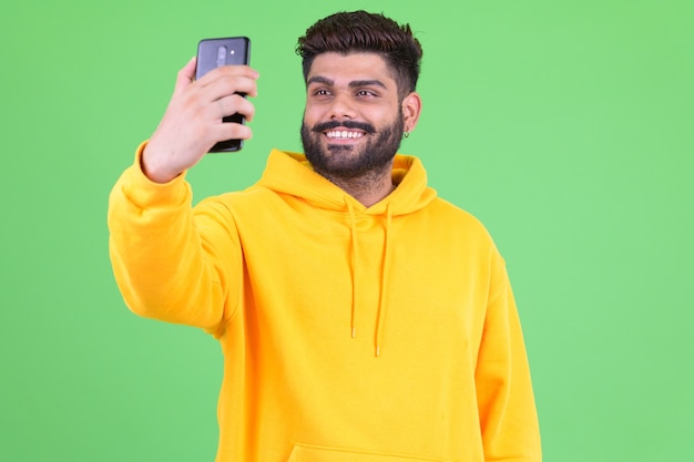Portrait of young overweight bearded Indian man wearing hoodie against chroma key with green wall