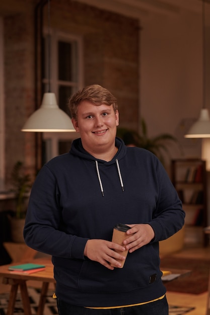 Portrait of young overweight businessman in casual clothing drinking coffee and smiling at front standing at office