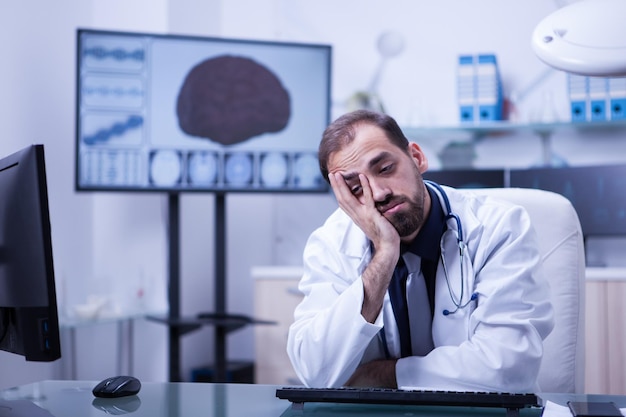 Portrait of young and overworked doctor with a brain image behind it in hospital cabinet. Fatigued doctor after long hours working in hospital.