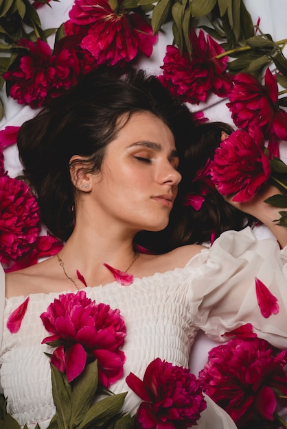 Portrait of a young, peaceful and gentle girl lying surrounded by scattered flowers, peonies. Top view