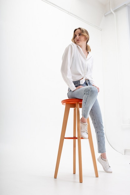 Portrait of young pensive caucasian woman posing in shirt, blue jeans, sitting on stool