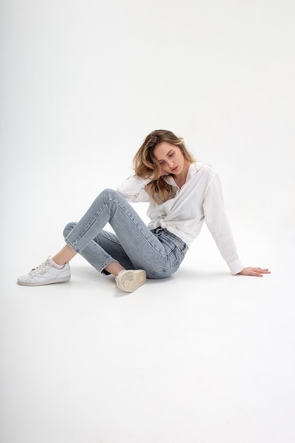 Portrait of young pensive caucasian woman posing in shirt and blue jeans, sitting on white floor. model tests of pretty girl in basic clothes on cyclorama