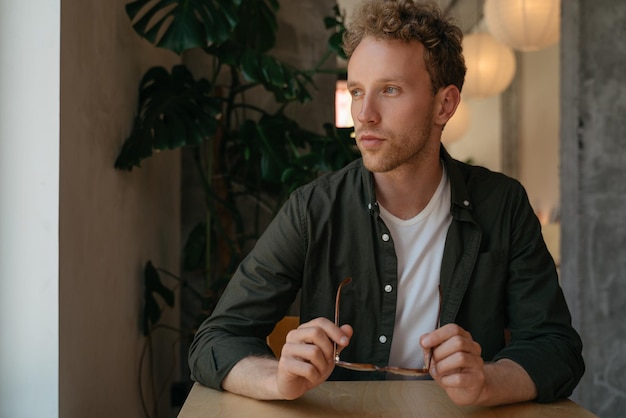 Photo portrait of young pensive man holding eyeglasses planning project looking at window sitting in cafe