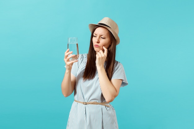 Portrait of young pensive woman in blue dress, hat holding and drinking clear fresh pure water from glass isolated on blue background. Healthy lifestyle, people sincere emotions concept. Copy space.