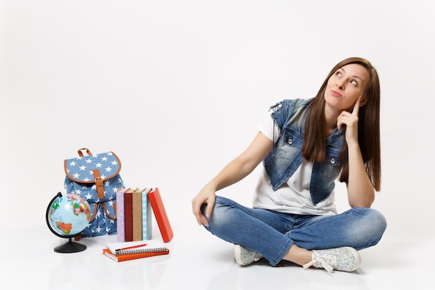 Ritratto di giovane studentessa pensierosa in abiti di jeans che guarda in alto sognando seduta vicino a libri scolastici zaino globo isolati