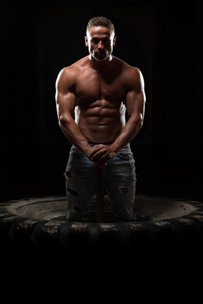 Photo portrait of a young physically fit man workout at gym with hammer  muscular athletic bodybuilder fitness model posing after exercises on tractor tire