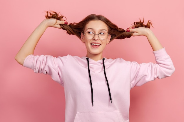 Портрет молодой игривой брюнетки в круглых очках, делающей хвостик с длинными каштановыми волосами и улыбающейся, дурачящейся или флиртующей. Съемка в помещении на розовом фоне
