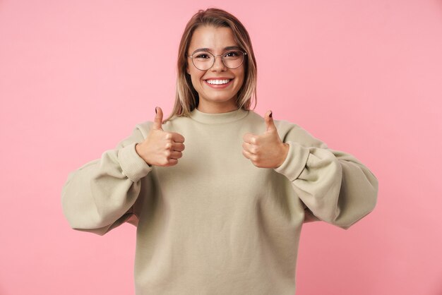 Portrait of young pleased woman in eyeglasses showing thumbs up and smiling isolated on pink