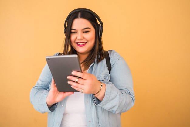 Ritratto di giovane donna plus size ascoltando musica con cuffie e tavoletta digitale all'aperto su giallo.