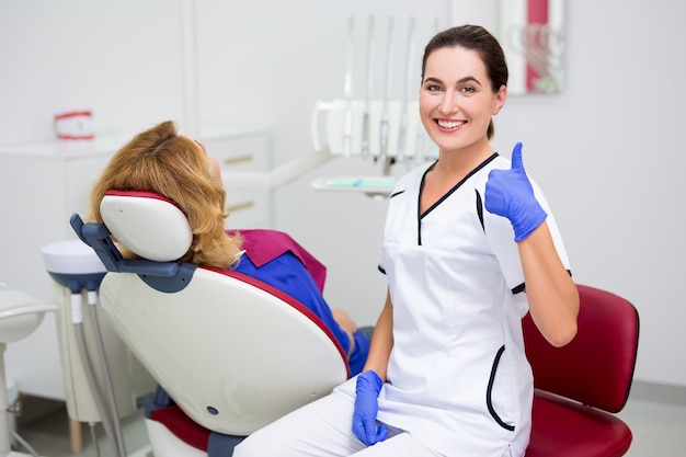 若いポジティブな女性の歯科医の親指を立てて、歯科用椅子に座っている患者の肖像画