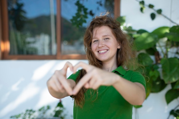 Portrait of a young positive girl next to her house shows a heart with her hands eco concept country