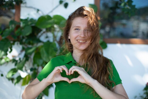 Portrait of a young positive girl next to her house shows a heart with her hands eco concept country