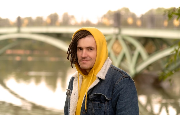 Photo portrait of young positive man with dreadlocks in hood posing in city park on background of blurred bridge in cloudy weather