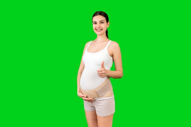 Portrait of young pregnant woman wearing bandage on her belly and showing thumb up sign at green background with copy space. Orthopedic abdominal support belt concept.