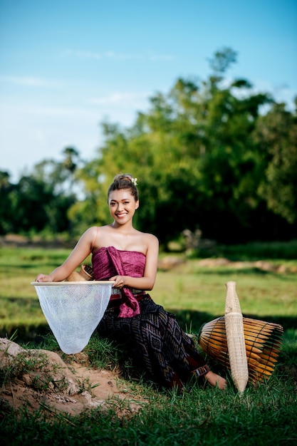 Portrait young pretty asian woman in beautiful thai traditional\
clothes at rice field, she sitting near fishing equipment