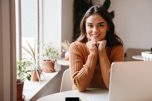 ラップトップコンピューターを使用して屋内のカフェに座っている若いかなり美しい女性の肖像画。