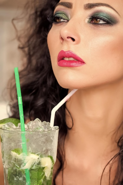 Portrait of young pretty brunette woman with curly hair and bright makeup with alcoholic mojito cocktail from mint soda light rum ice cubes and lime with straw vertical picture